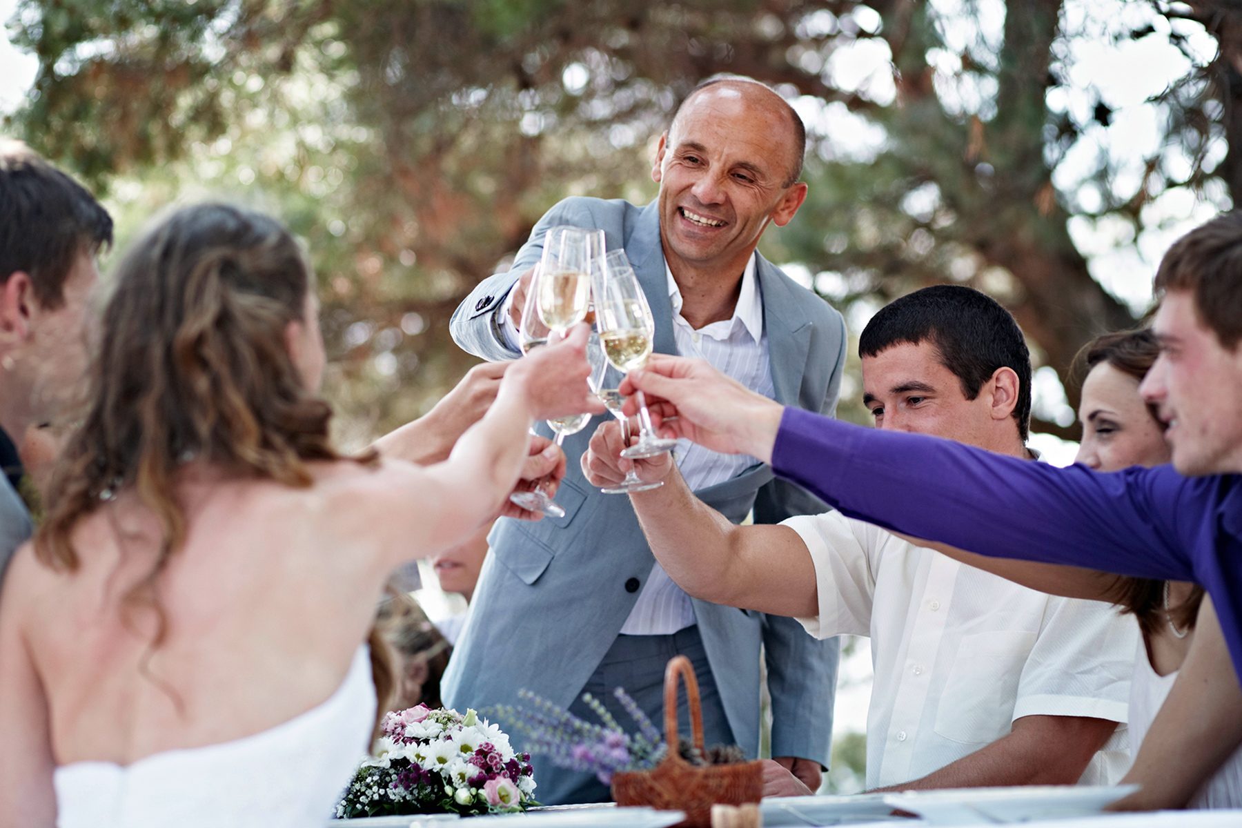 A group of people are sitting around a table outdoors, toasting with champagne glasses. They seem to be enjoying a celebratory moment. The table has a flower arrangement and wicker basket. Trees are visible in the background, providing a lush setting.