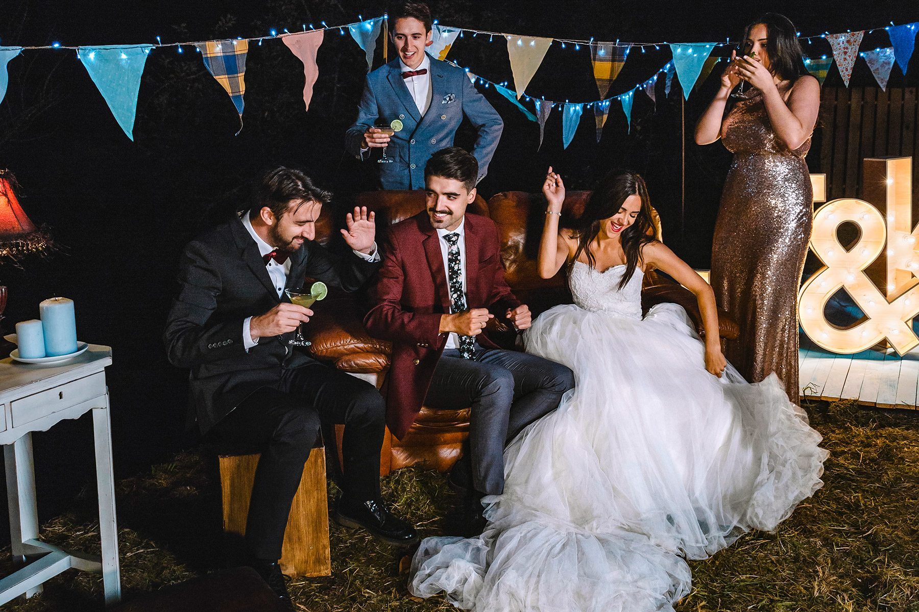 Happy wedding couple having fun sitting on sofa with their friends on a night field party