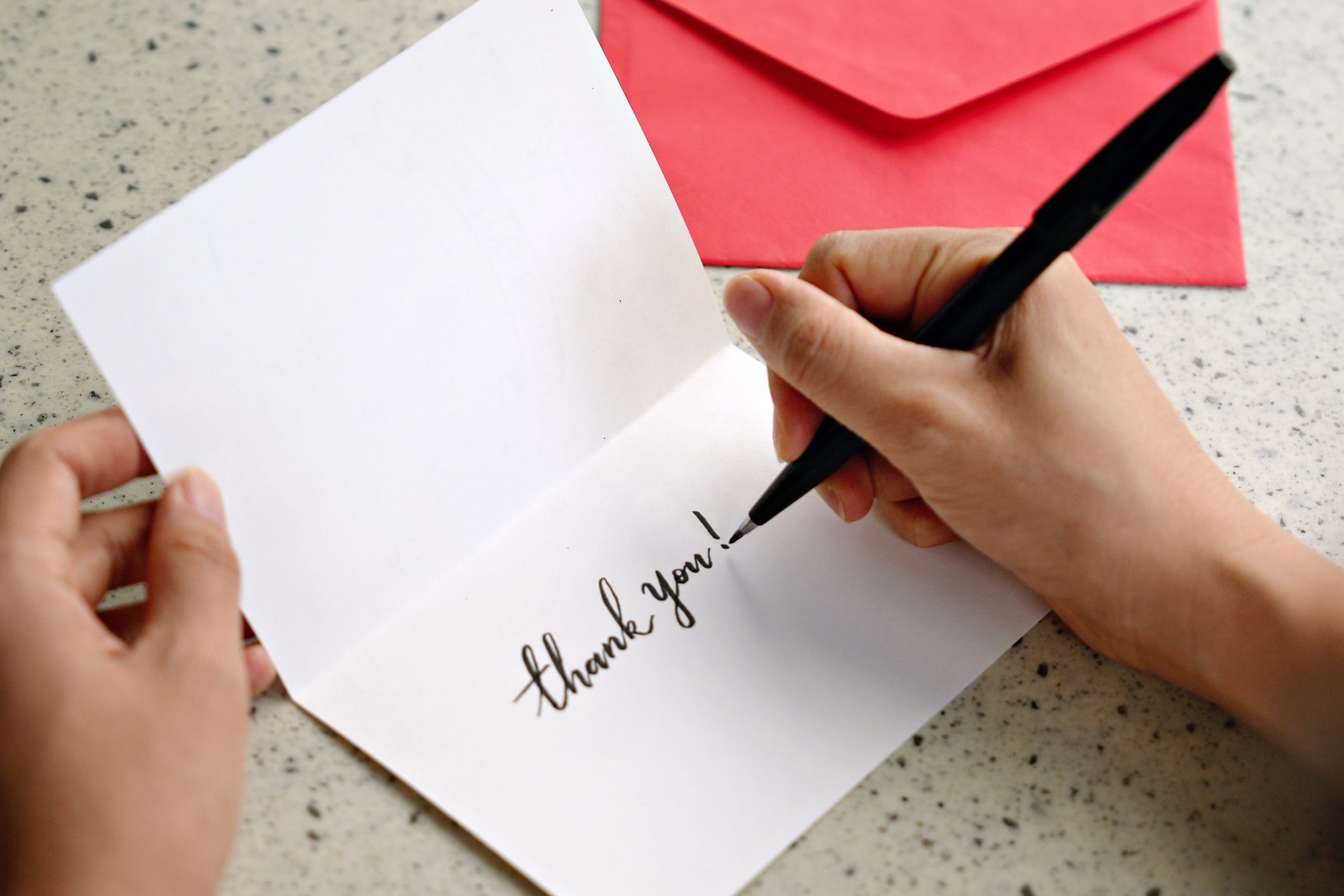 With a red envelope nearby, a woman's hands write a "Thank You" note inside a greeting card.