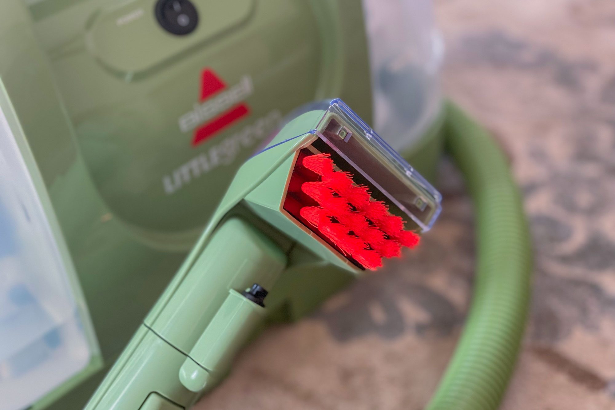 A cleaning machine with a red-bristled brush attachment rests on a patterned surface. Brand name "Bissell Little Green" visible on the machine.