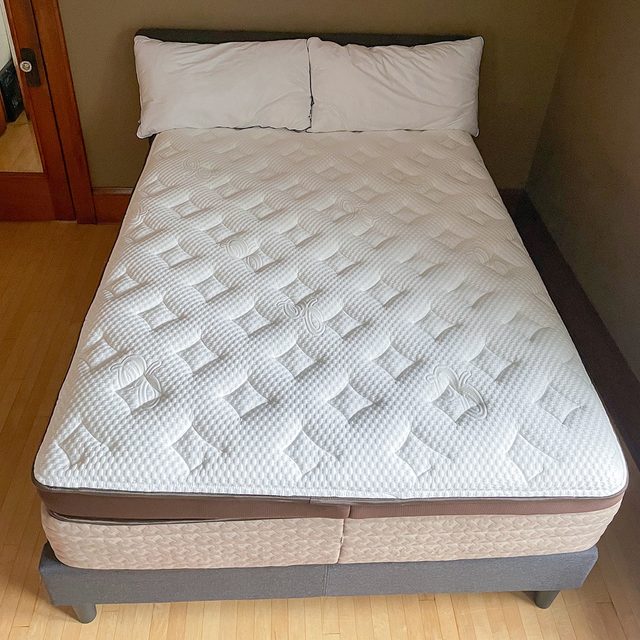 Bed with white, quilted mattress and two pillows sits in a simple room with hardwood floors and a wooden door.