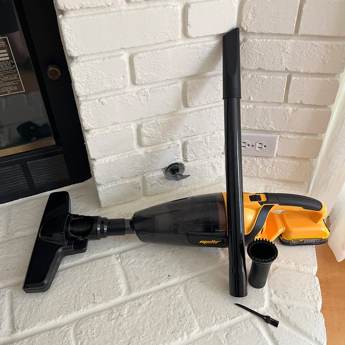 Cordless vacuum positioned on white brick fireplace, surrounded by attachments; black nozzle and brush, with a black and yellow color scheme.