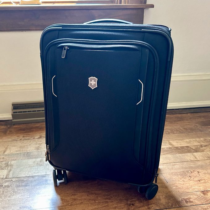 A black suitcase stands on wooden floor, featuring four wheels and a silver emblem. Window light creates shadows in the room.