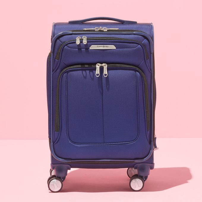 Suitcase stands upright on four wheels, featuring multiple zippered compartments, against a pink background. 