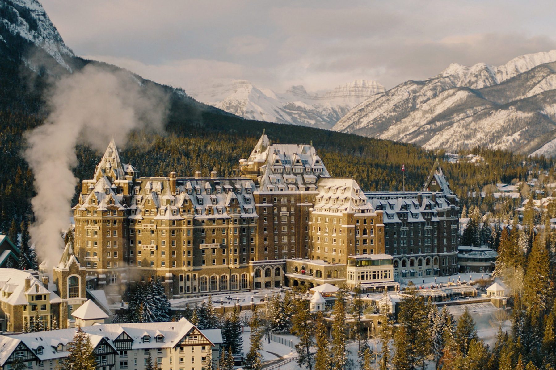 Grand hotel stands surrounded by snow-covered forest, with steam rising, nestled in mountainous landscape under a cloudy sky.