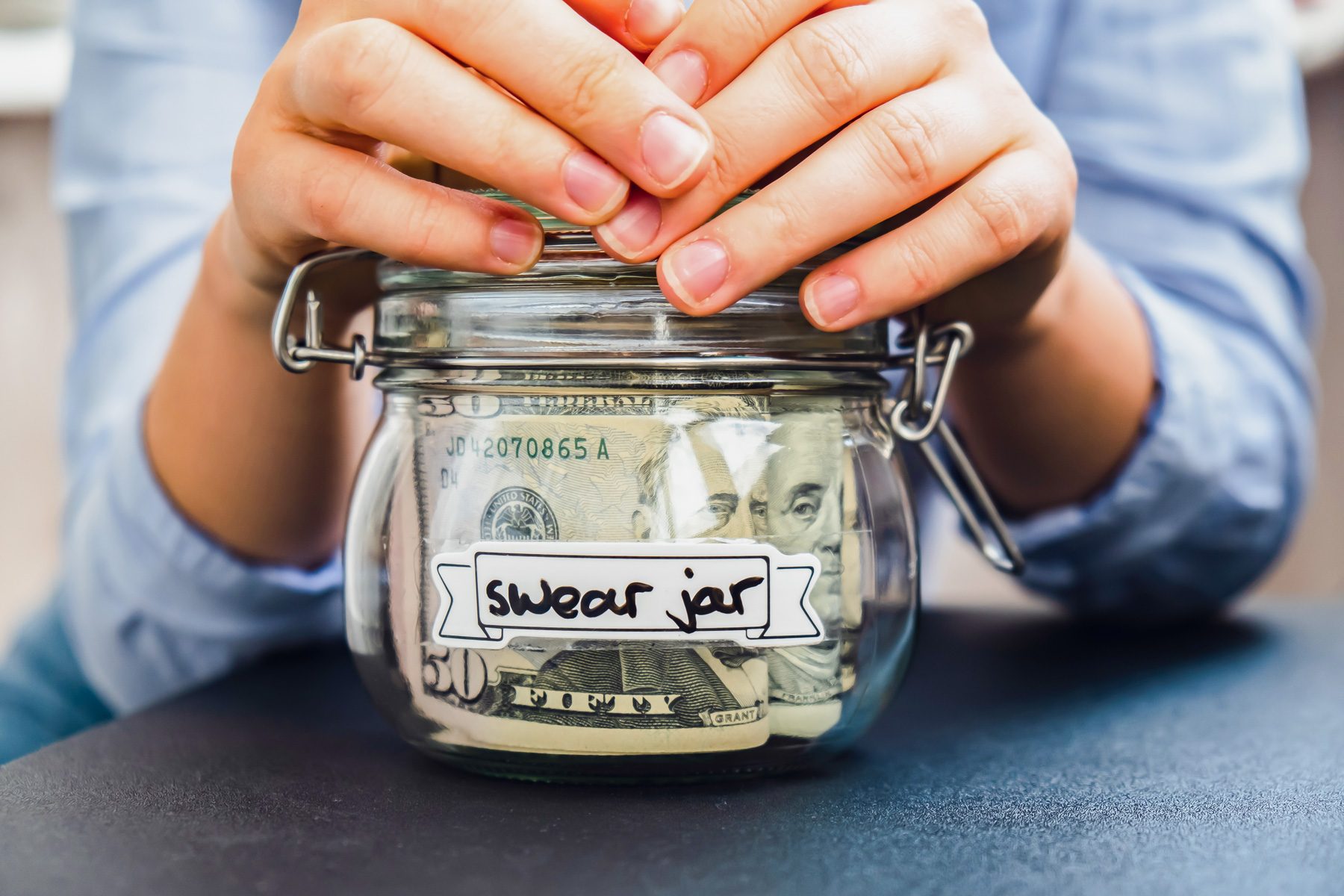 Hands hold a glass jar labeled "swear jar," filled with US dollar bills, on a table.