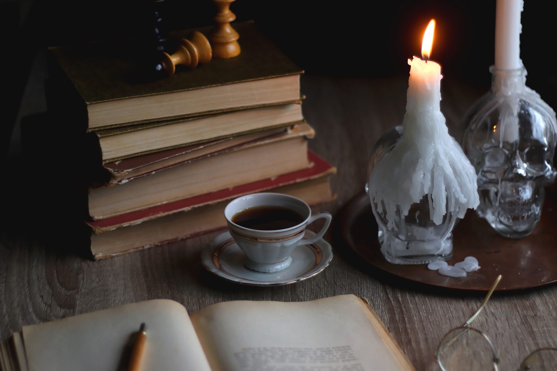 A stack of books sits beside a lit candle on a skull-shaped holder, with coffee, an open book, and glasses on a wooden table.
