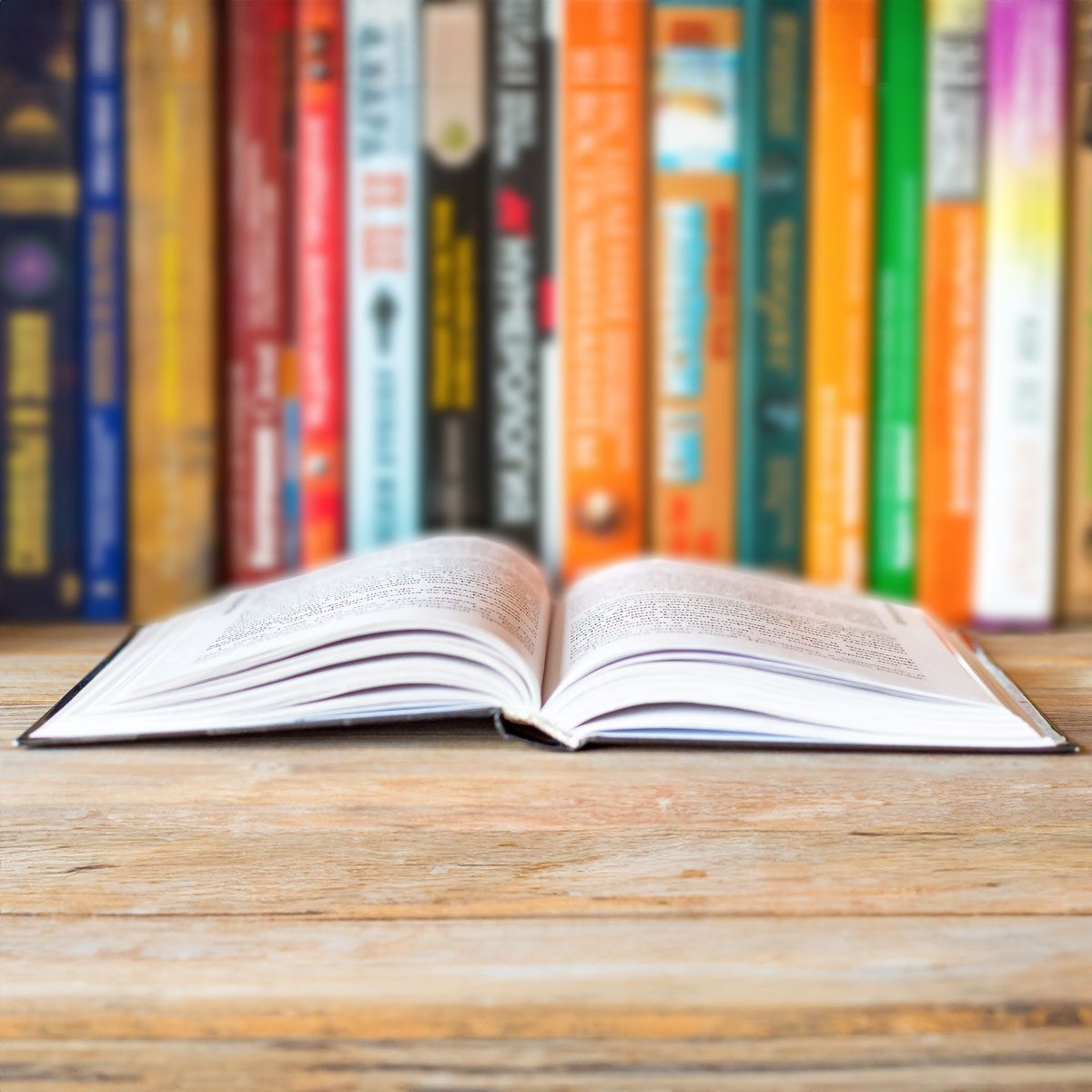 An open book rests on a wooden table, surrounded by a colorful, blurred bookshelf background.