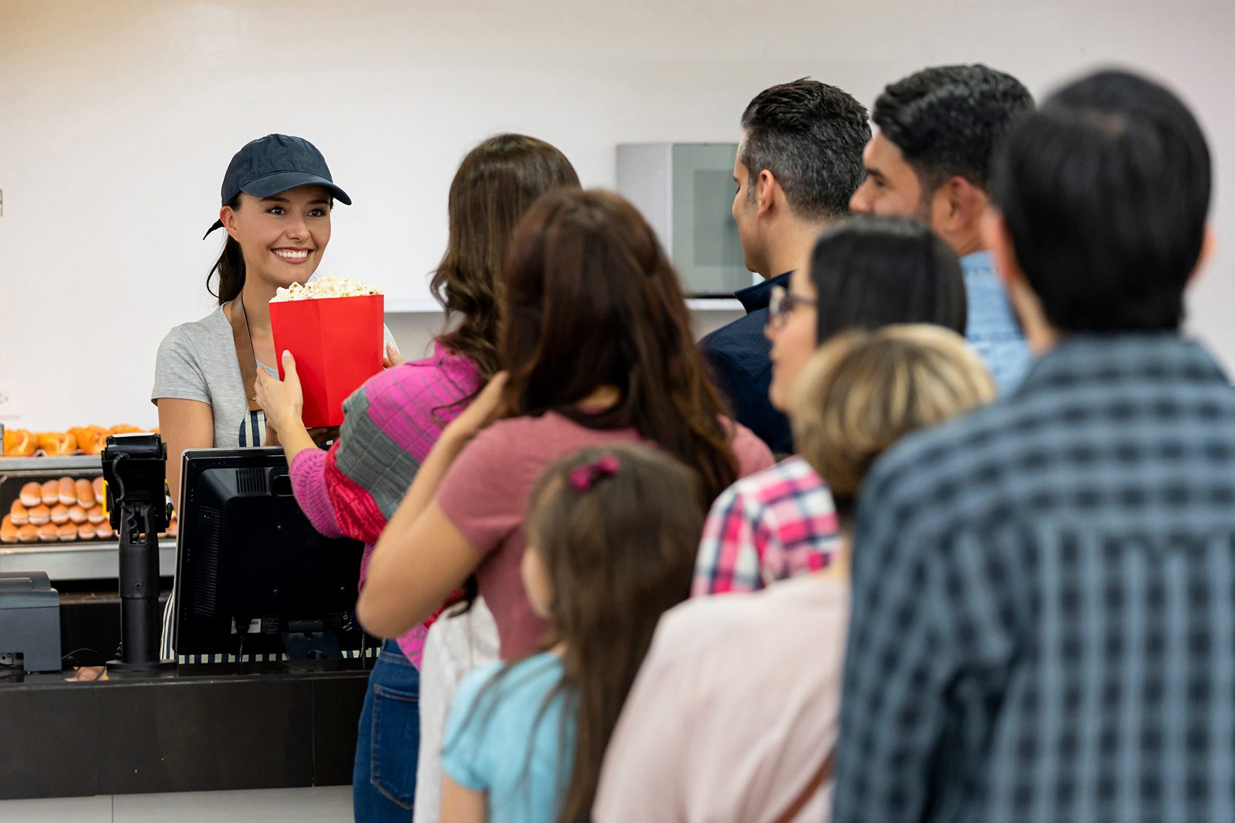 Cashier hands popcorn to customer in a concession line, surrounded by people and food items in the background.