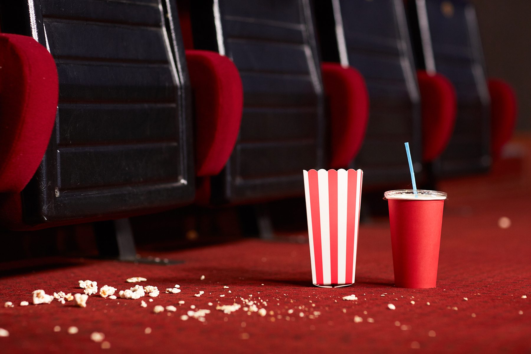 Empty popcorn box and drink cup rest on carpeted theater floor, surrounded by scattered popcorn near red cushioned seats.