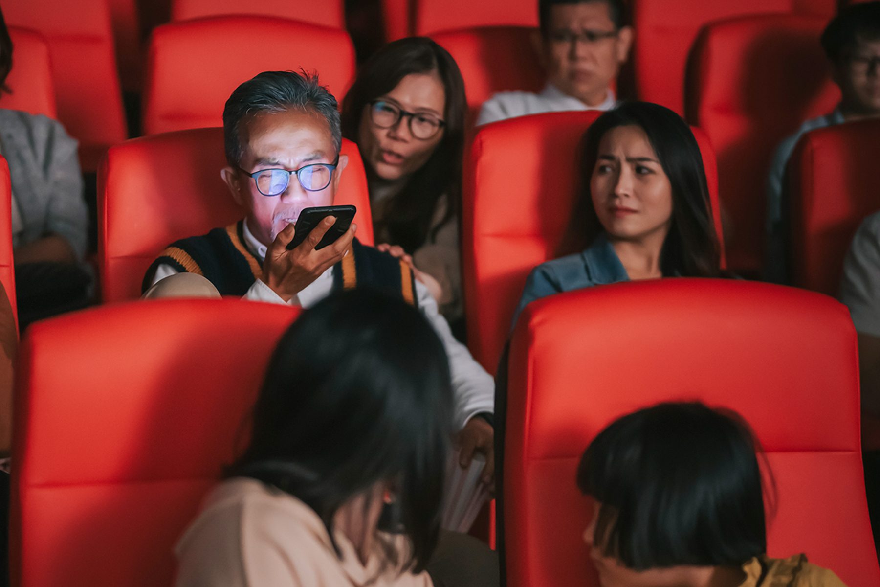 Person uses phone in a dimly lit cinema, while others seated nearby appear annoyed or distracted.