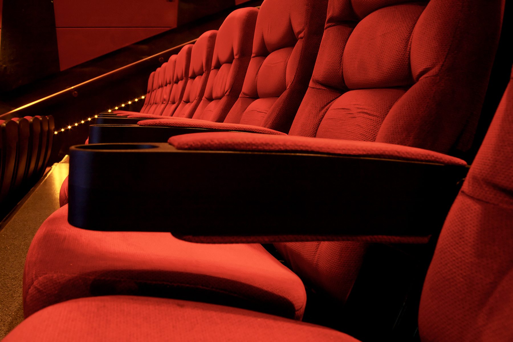 Red theater seats lined up, unoccupied, in a dimly lit auditorium with glowing aisle lights.