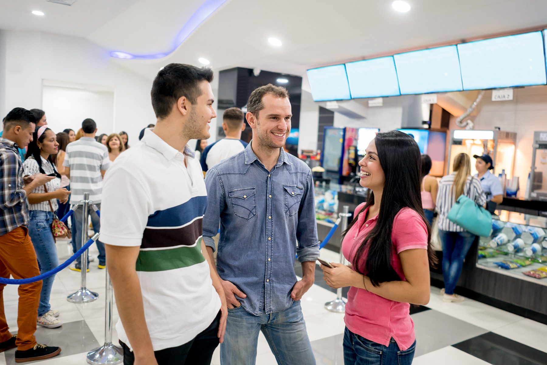 Three people are chatting happily in a movie theater lobby, surrounded by others waiting in line near the concession stand.