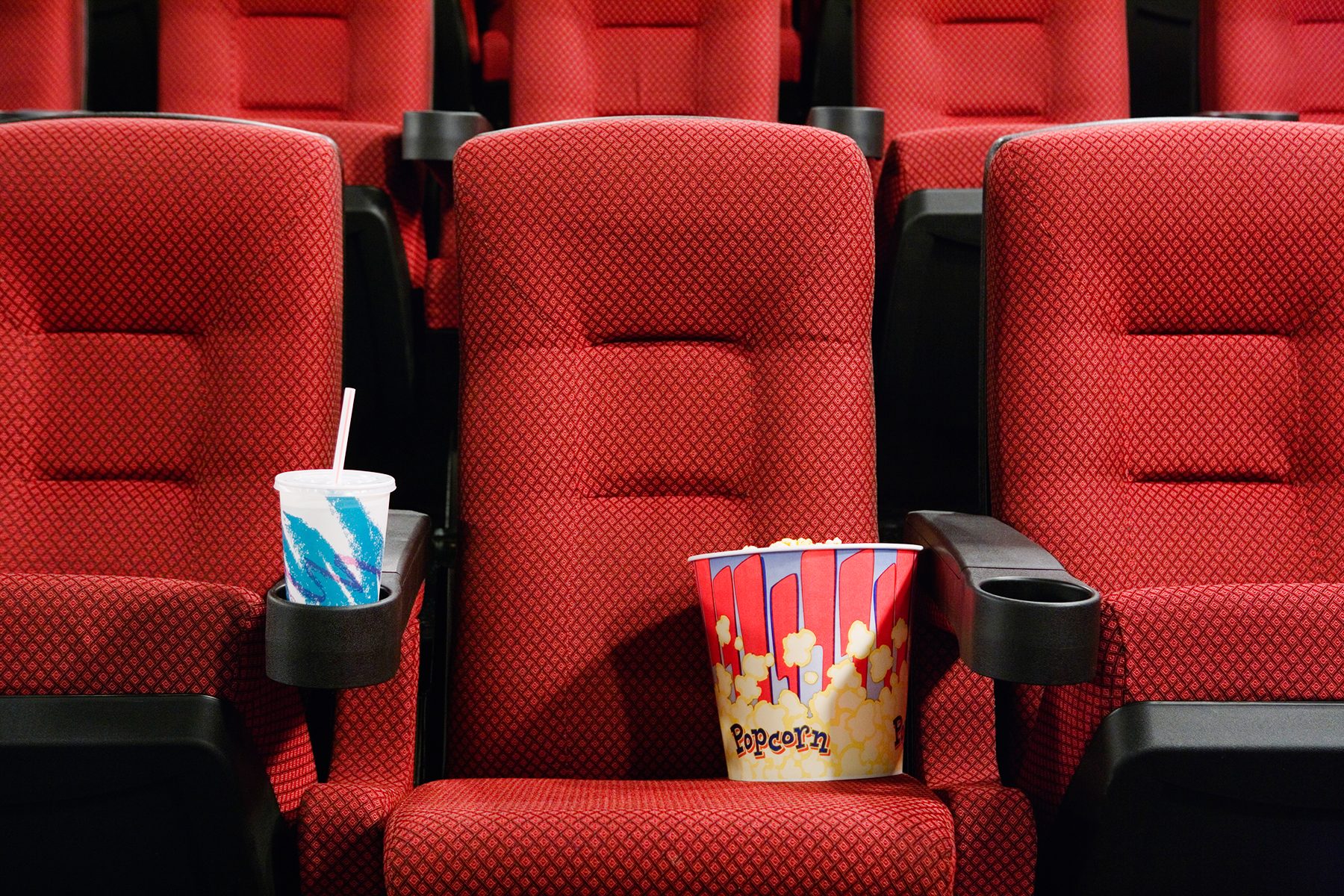 Red theater seats hold a drink and popcorn. The environment is an empty cinema, creating a waiting atmosphere.