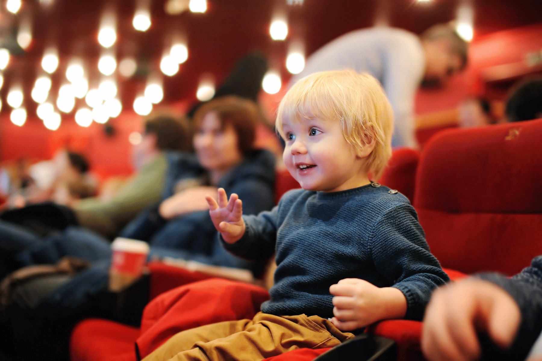 Child sits excitedly in a theater, surrounded by plush red seats and blurred audience members, under bright round lights.
