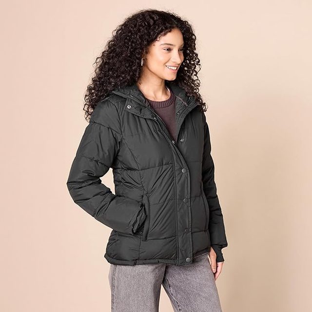 A person stands smiling, wearing a dark puffer jacket. The background is a plain beige color.