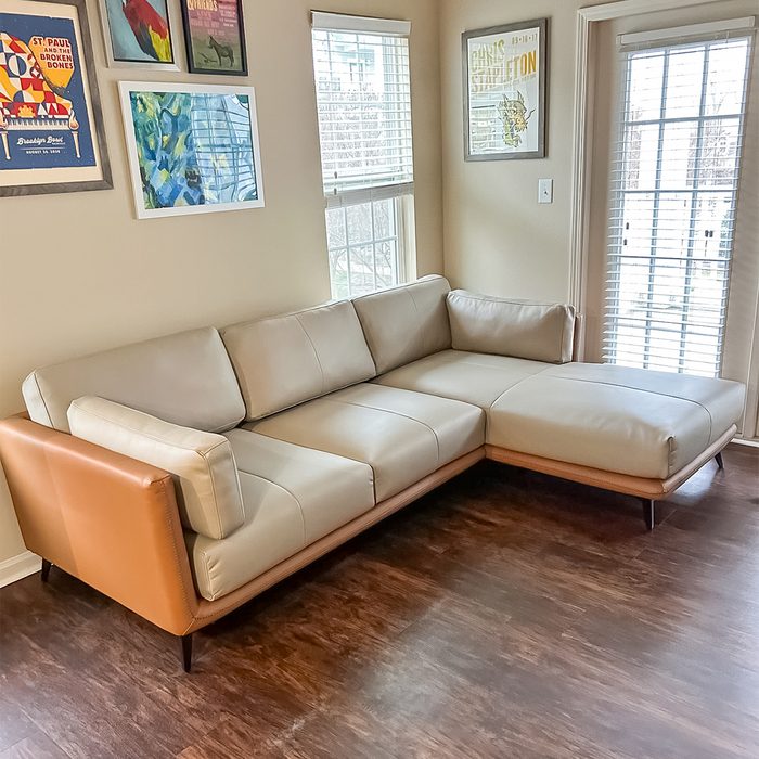 A light brown L-shaped sofa sits in a living room corner, surrounded by windows with blinds and colorful framed wall art.
