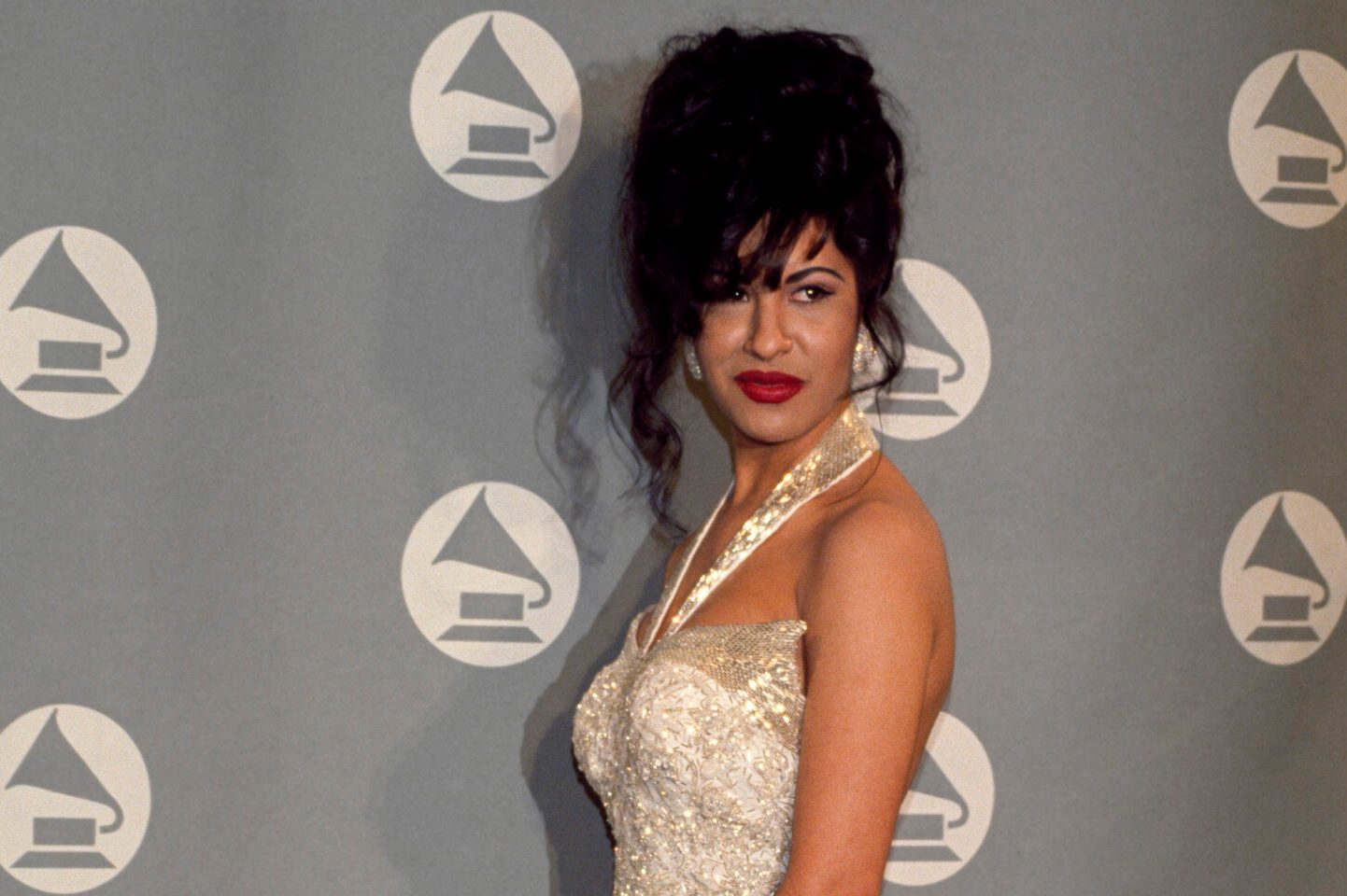A woman in a white dress poses on a red carpet, with a backdrop featuring repeated Grammy award logos.