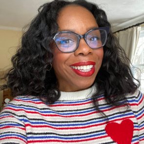 Person smiling, wearing glasses and a striped sweater with a red heart, indoors with light walls and large windows in the background.