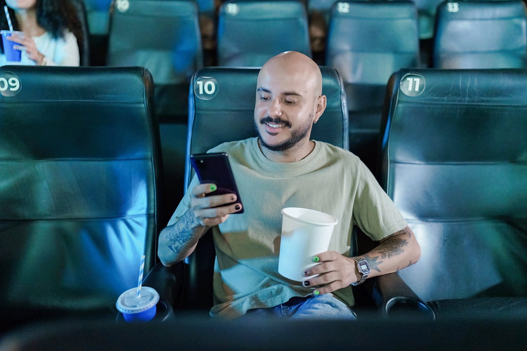 Person using phone sits in theater, holding popcorn. Surrounding empty seats are numbered 09, 10, 11, 12, 13. A drink rests beside them.
