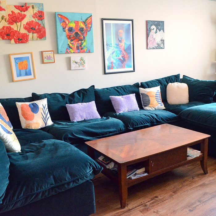 A dark green sectional sofa holds colorful pillows, set against an art-filled wall, with a wooden coffee table in front, on a wooden floor.
