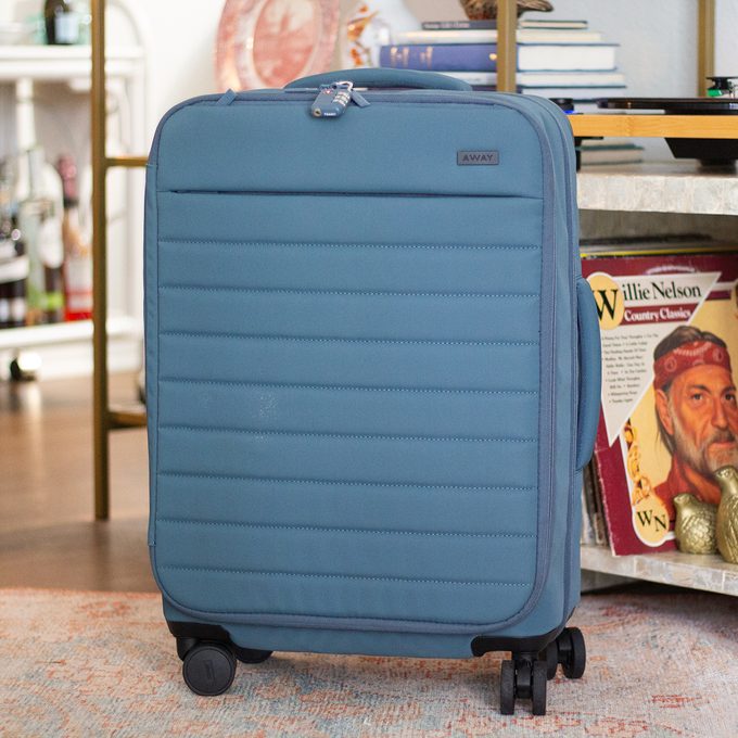Blue suitcase stands upright on four wheels, placed in a cozy living room with a rug and bookshelf. Books and a magazine are nearby.