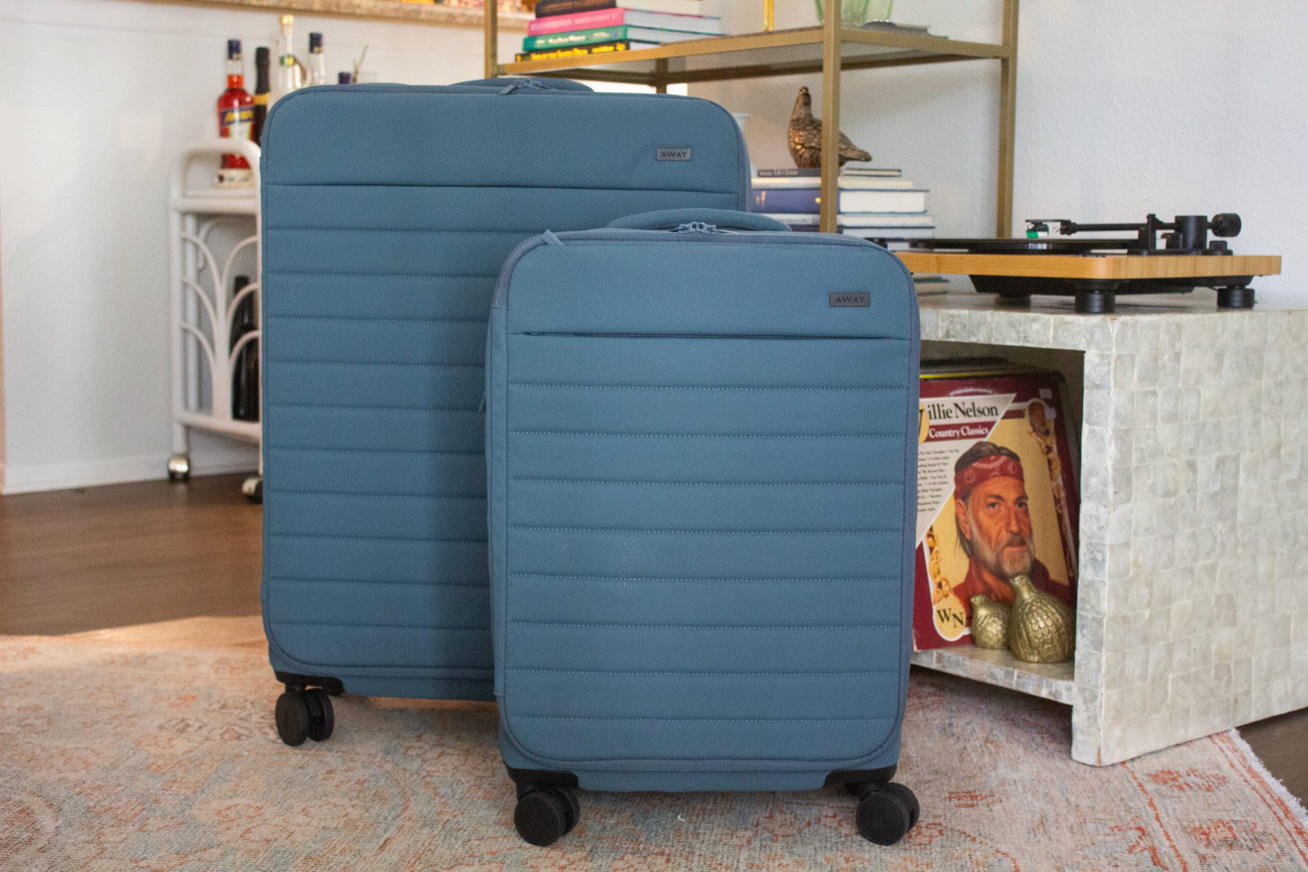 Two blue suitcases stand upright on wheels, indoors near a vintage record player and shelf with books and decor, in a cozy room.