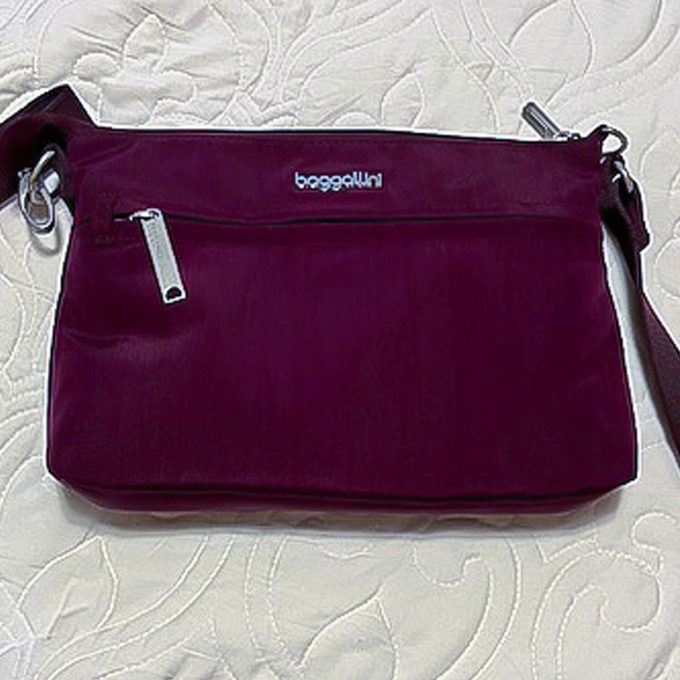 Maroon bag resting on a quilted white surface. It features a zipper and brand name 