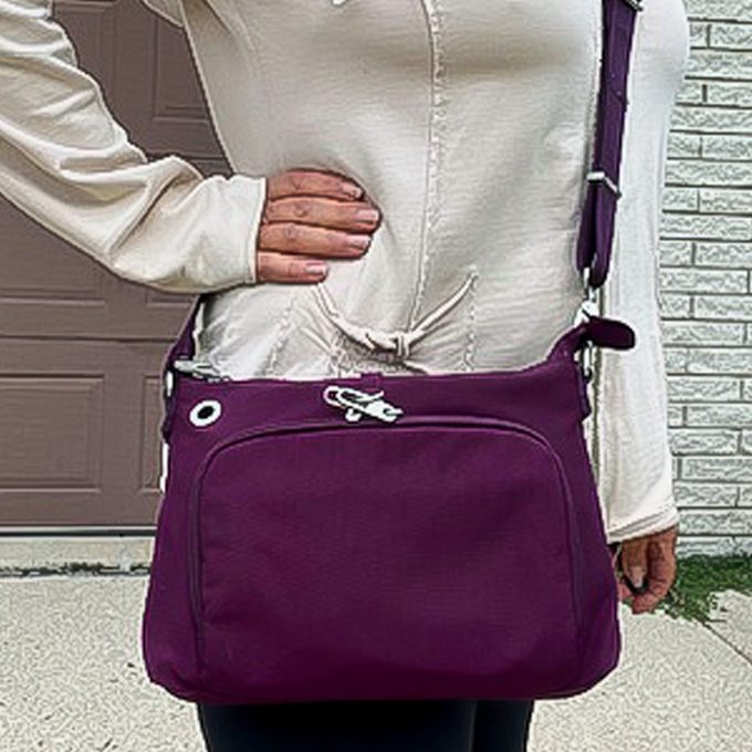 A person carries a purple crossbody bag in front of a garage and brick wall, wearing a cream top.