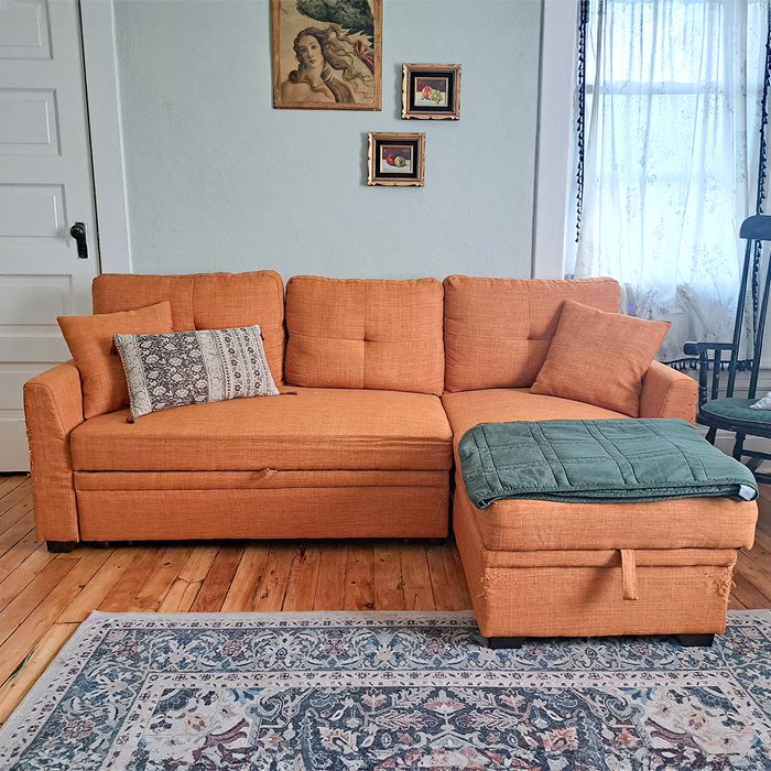 Orange sectional sofa sits in a cozy living room, adorned with patterned pillows, beneath framed art on a light blue wall near lace-curtained window.