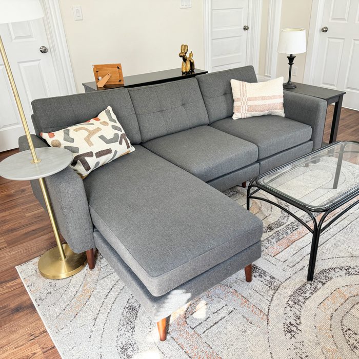 Gray sectional sofa sits in cozy living room, accompanied by a glass coffee table, decorative pillows, and a modern floor lamp.