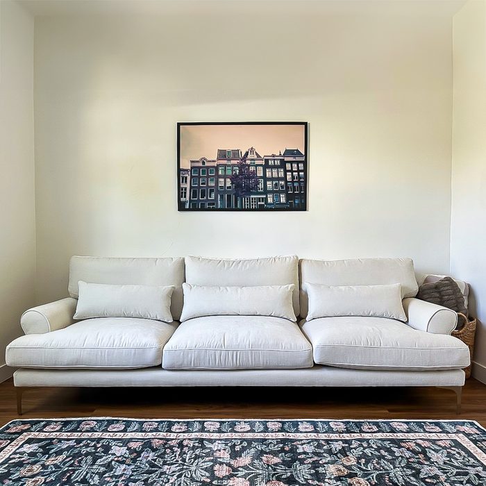 A cream-colored sofa sits against a wall beneath a framed cityscape photo, situated on a hardwood floor with a floral-patterned rug.