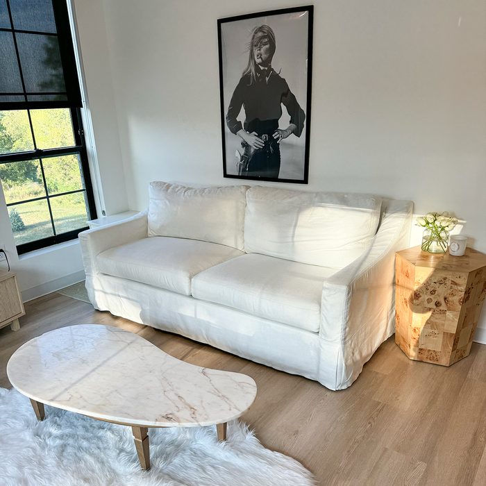 White couch sits in sunlit room, flanked by unique wooden table, beneath a black-and-white portrait. Nearby, a marble coffee table on a fluffy rug.