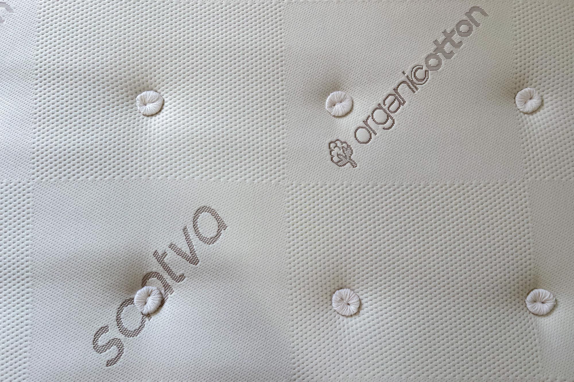 Mattress surface, displaying "saatva" and "organic cotton." Features tufted buttons across a textured, grid-patterned fabric.