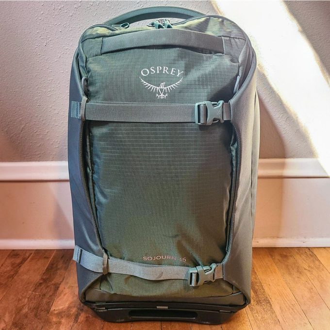 A green backpack stands upright on a wooden floor against a white wall, labeled 