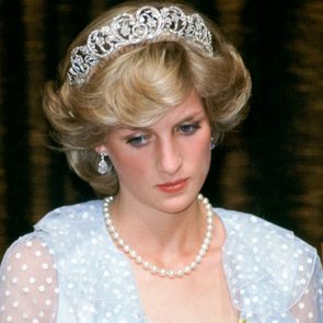 A woman with styled blonde hair wears a tiara and pearl necklace, looking down thoughtfully in a formal setting.