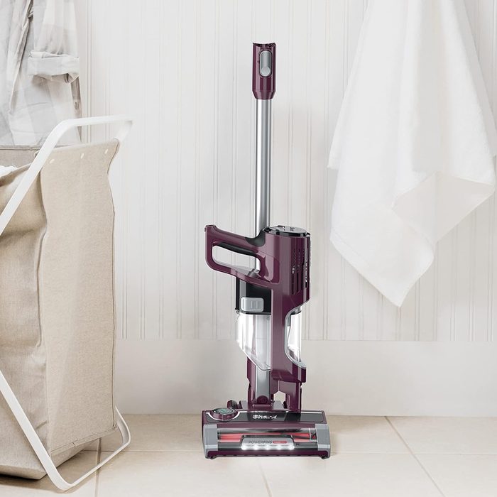 A vacuum cleaner stands upright on a tiled floor, next to a laundry basket and a hanging towel on a white paneled wall.