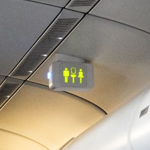 Ceiling sign illuminates restroom symbols beside an indicator light; positioned above airplane cabin seating with overhead compartments.