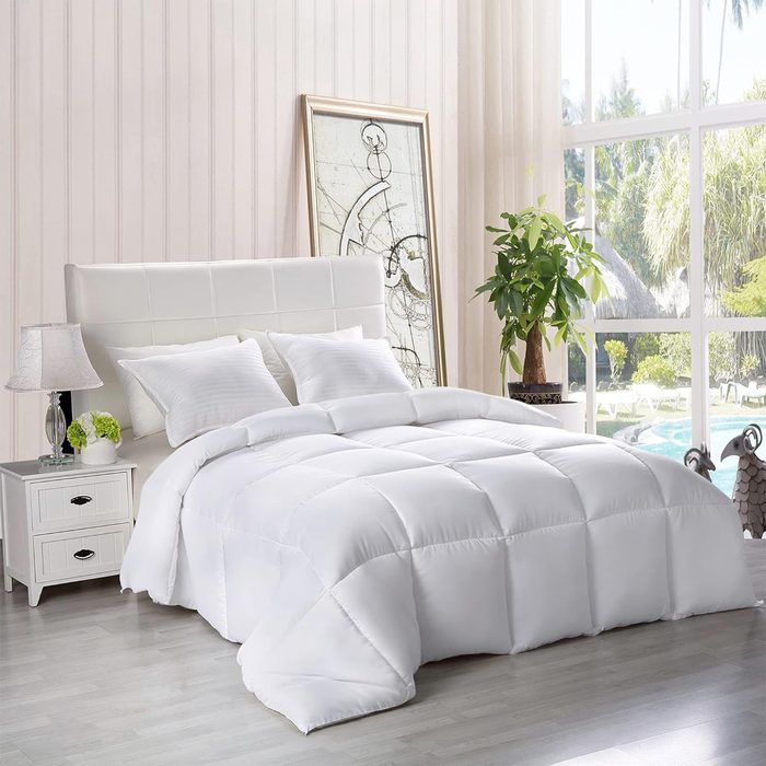 Bed with white quilted comforter sits in bright room, accompanied by nightstand, lamp, framed artwork, and large window overlooking trees and pool.