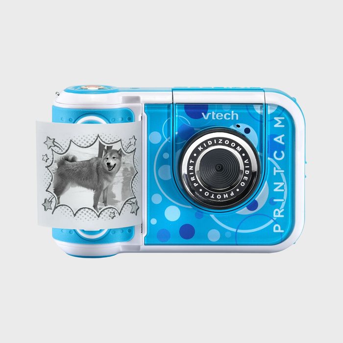 A blue children's camera prints a black-and-white dog photo with star decorations. The device features include VTech branding and 