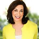 A person smiling, wearing a yellow top and standing in front of a blurred outdoor background featuring greenery.