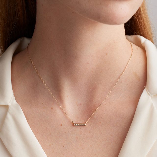 Necklace rests on a woman's neck, featuring a small horizontal bar pendant. She wears a cream blouse, providing a soft backdrop.