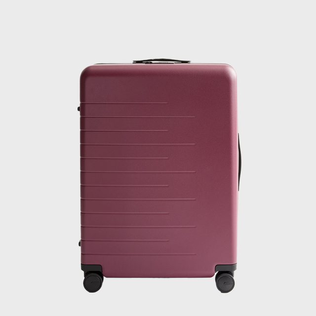 A burgundy suitcase stands upright with wheels, showcasing a smooth design, in a minimal white background.