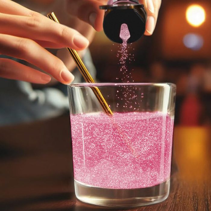 Glass with pink drink, hand sprinkles powder, golden stirrer inside, dimly lit room.