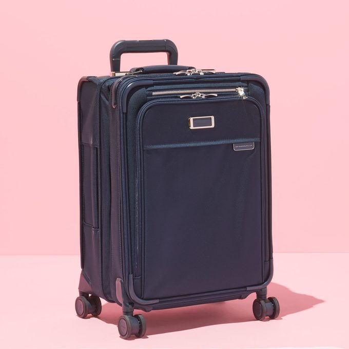 A black suitcase stands upright, stationary on its four wheels, set against a plain pink background.