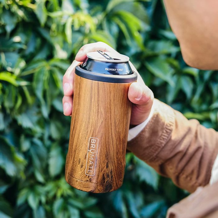 A hand holds a wooden-patterned BrÃ¼Mate can cooler in front of lush green leaves, showing the text 