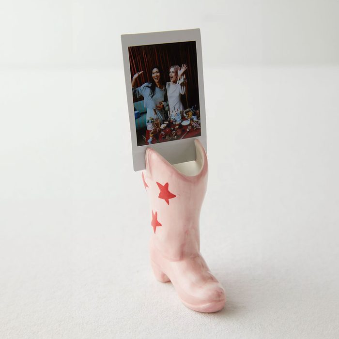 A pink ceramic boot with red stars holds a polaroid photo, set against a plain, light background.
