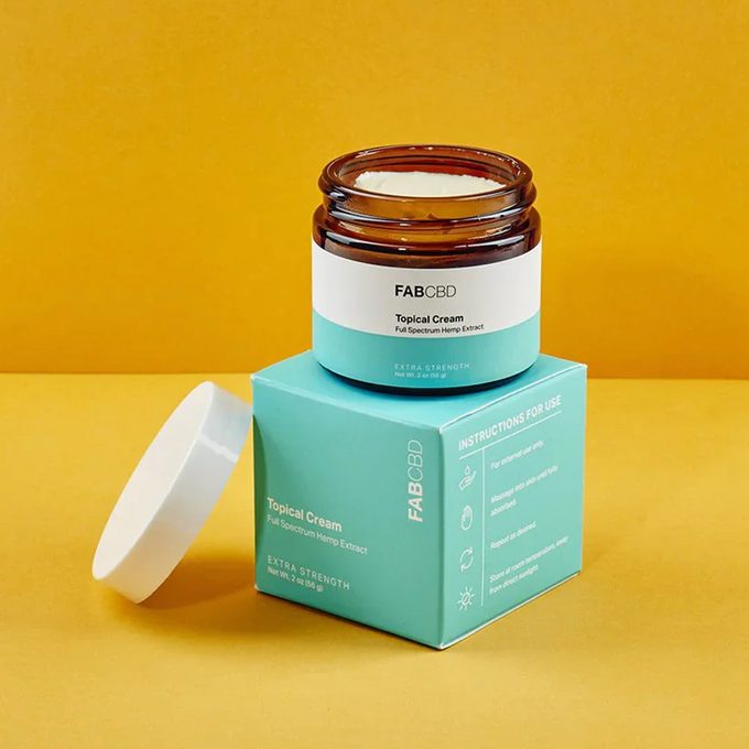 Jar of topical cream sits open on a teal box, against a yellow background. Text includes 