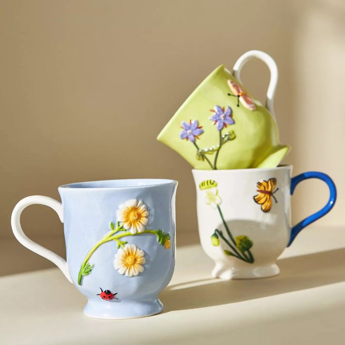 Ceramic mugs stand, featuring floral and insect motifs, against a softly lit beige background.