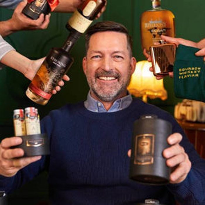 Man smiling, surrounded by outstretched hands holding whiskey bottles and a flask; indoor setting with warm lighting and a lamp in the background.